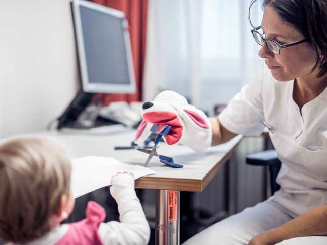 Ärztin mit Handpuppe erklärt einem Kind spielerisch ein Hilfsmittel zum Schneiden etwas